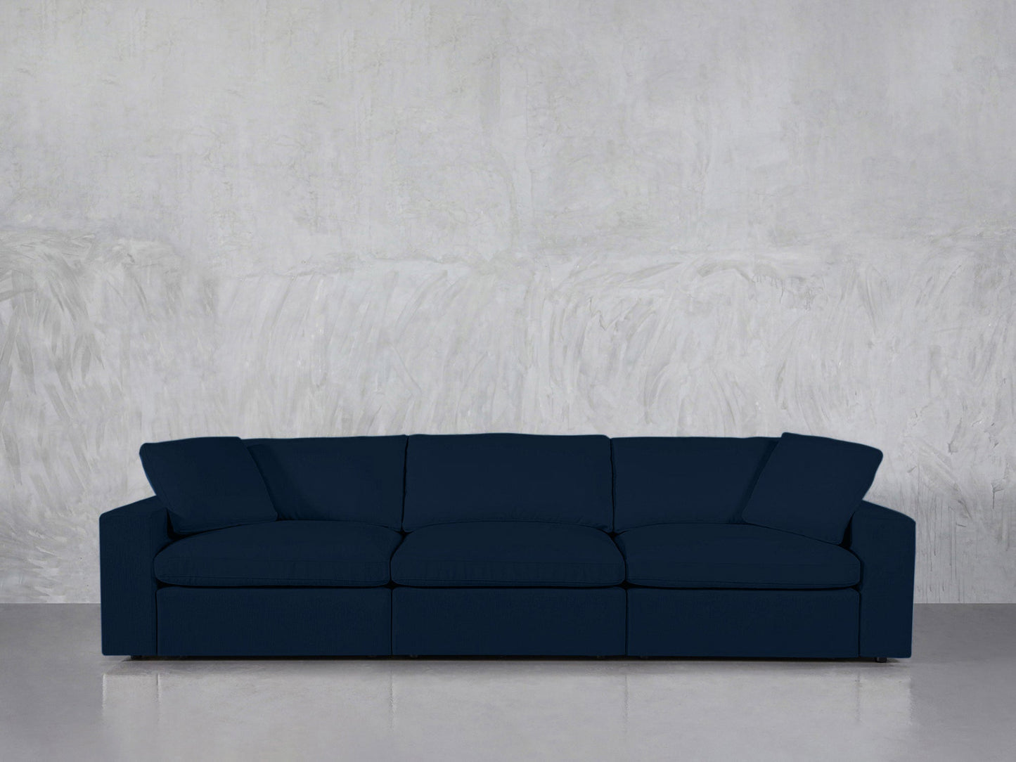 The 3-Seat Modular Sofa in dark blue, featuring three seat cushions and two angled armrests, is placed against a textured light gray wall on a concrete floor.
