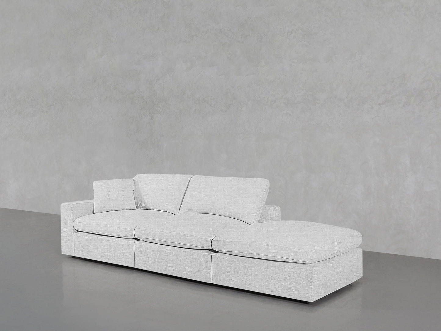 The 3-Seat Modular Lounger Sofa, in modern light gray, features three seats and a right-side chaise, positioned against a textured gray wall and smooth gray floor.
