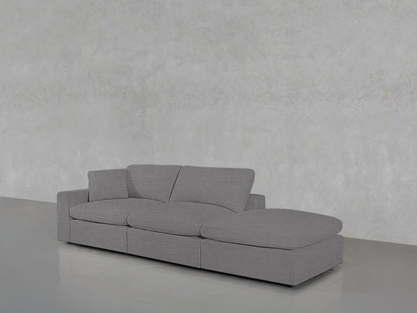 A 3-Seat Modular Lounger Sofa with plush gray cushions is placed on a glossy light gray floor against a textured light gray wall in a minimalist, empty room.