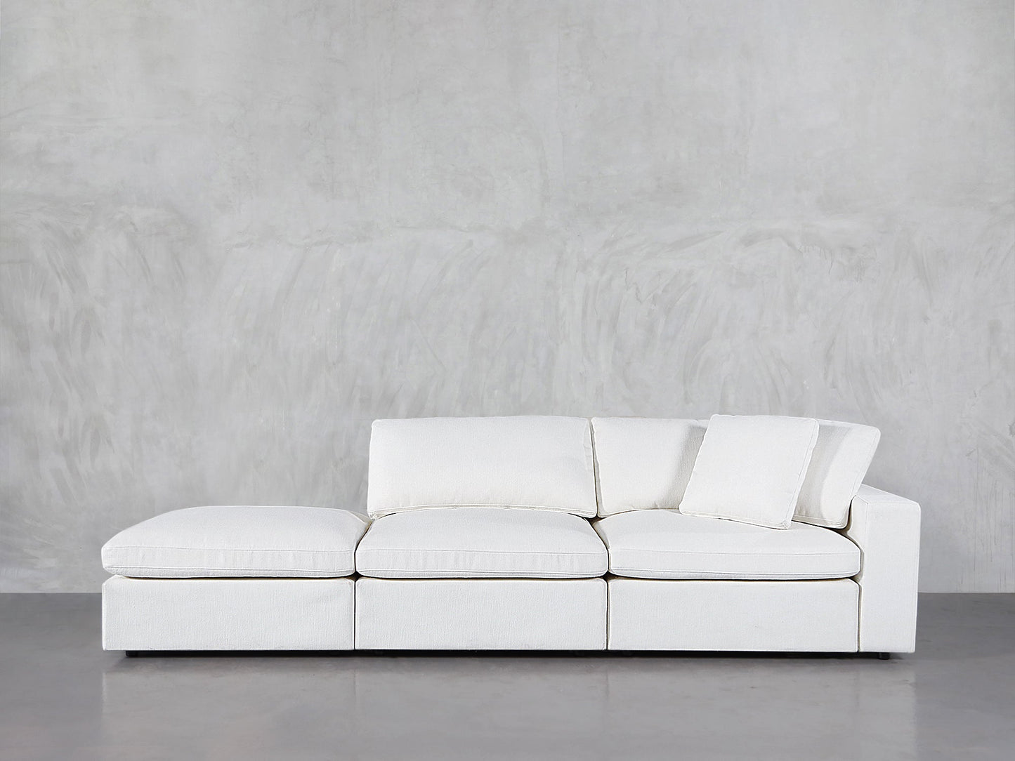 The 3-Seat Modular Lounger Sofa, upholstered in modern white with three seat and back cushions plus a matching throw pillow, sits against a light gray textured wall on a polished floor.