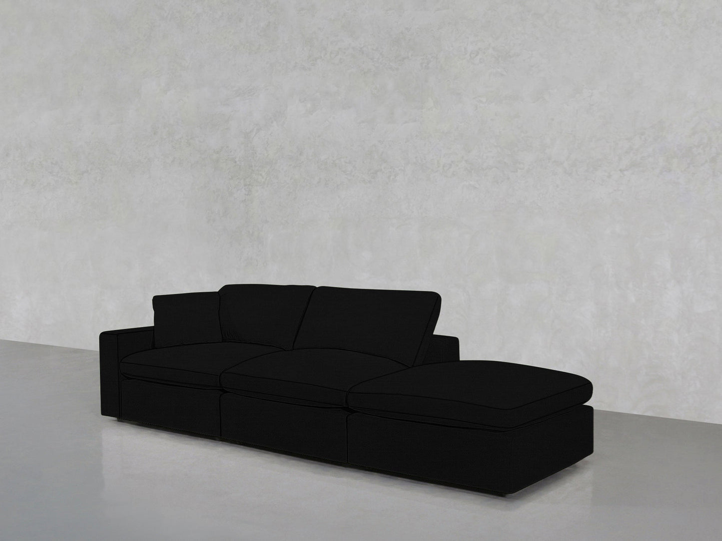 The 3-Seat Modular Lounger Sofa in black features an extended chaise and is displayed against a plain light gray textured wall with a smooth gray floor.