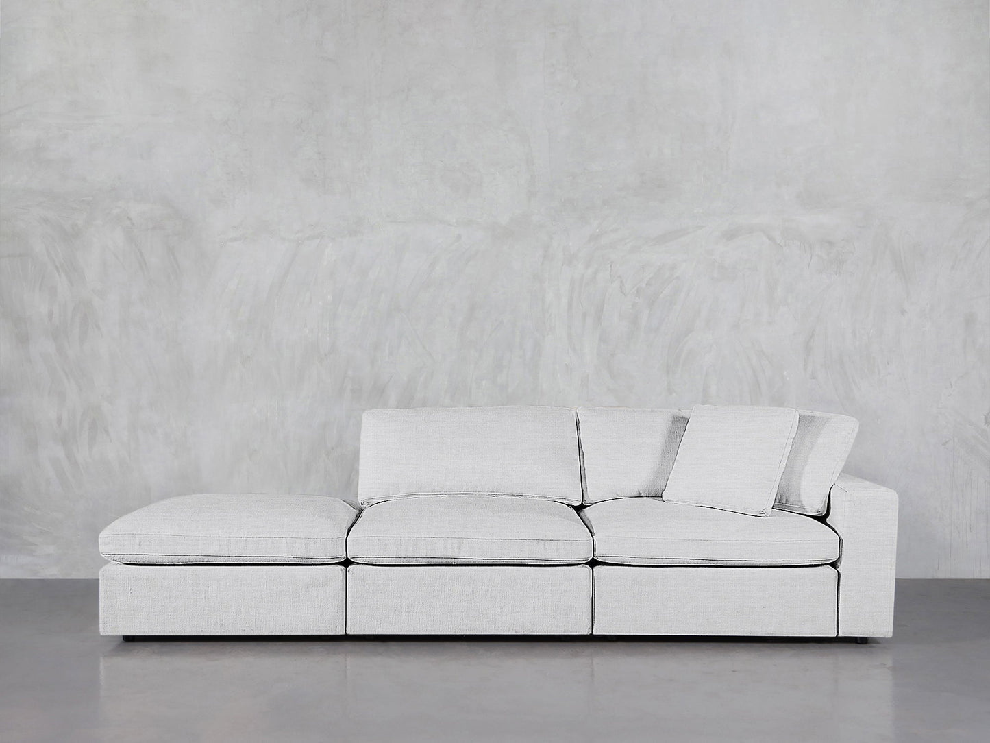 The 3-Seat Modular Lounger Sofa, featuring a modern design in light gray with square cushions and clean lines, is showcased against a plain, textured light gray wall and a smooth floor.
