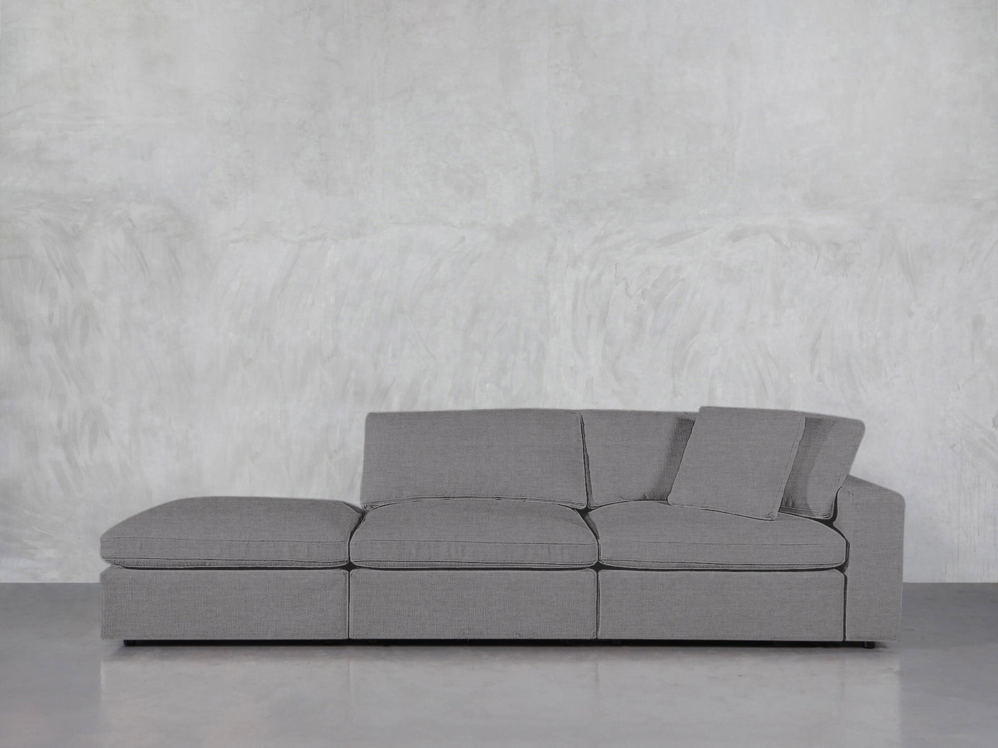The 3-Seat Modular Lounger Sofa features a modern gray design with three cushions and two angled back pillows, displayed against a light gray textured wall and polished floor.