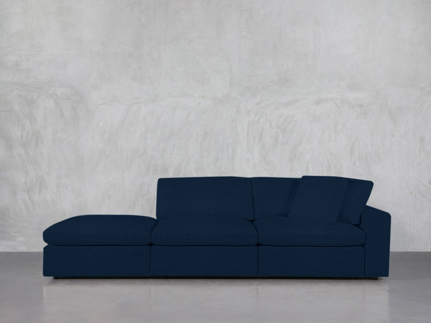 A 3-Seat Modular Lounger Sofa in dark blue with a chaise lounge is positioned against a light gray textured wall on a polished concrete floor.