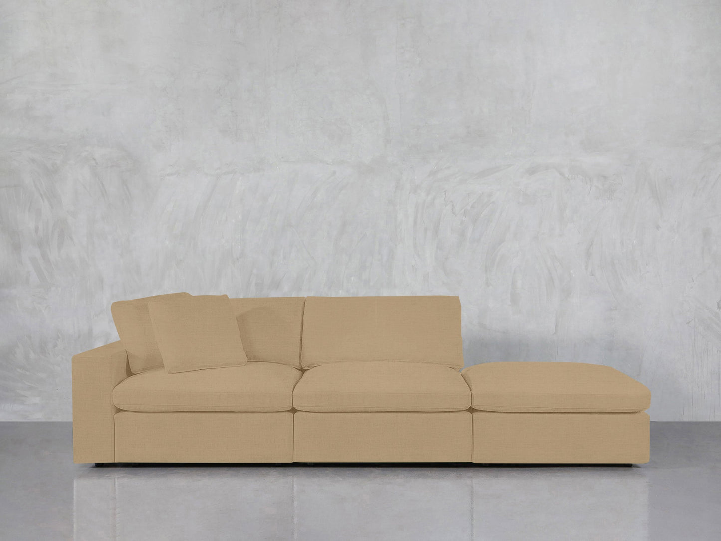 A 3-Seat Modular Lounger Sofa in beige, featuring an attached chaise lounge and two matching cushions on the left, is set against a light gray textured wall with a reflective floor beneath it.