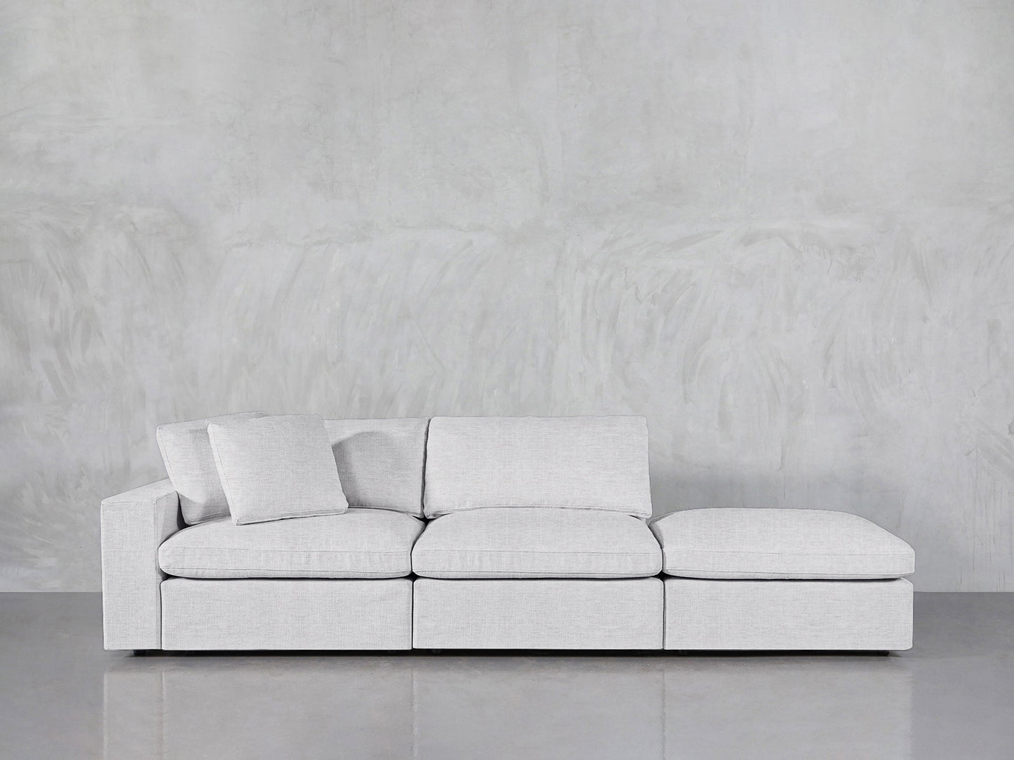 The 3-Seat Modular Lounger Sofa in modern light gray features three seat and back cushions plus two pillows, shown against a plain gray textured wall with a reflective floor.
