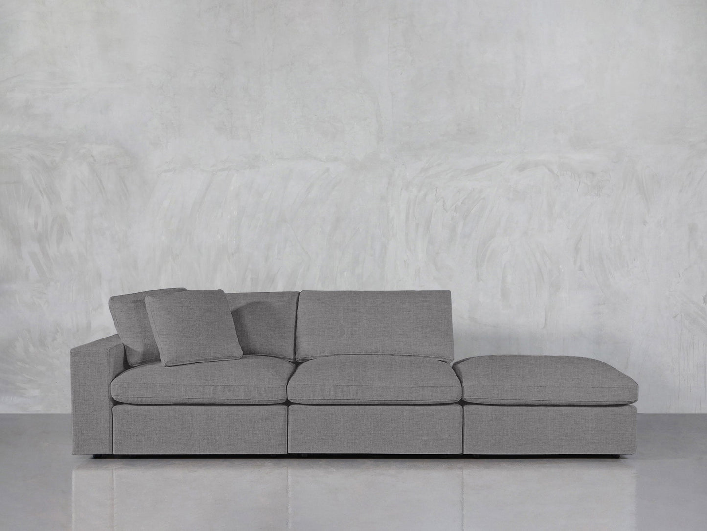 The 3-Seat Modular Lounger Sofa in modern light gray features three seat cushions, two matching pillows, and a right-side chaise lounge, set against a plain gray textured wall and shiny floor.