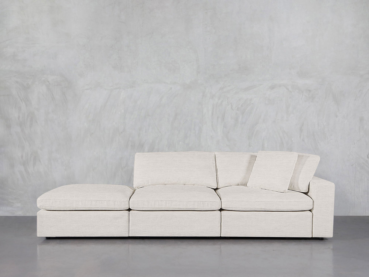 The 3-Seat Modular Lounger Sofa, in light beige with three seat cushions and two angled backrests, is placed against a textured gray wall on a polished concrete floor.