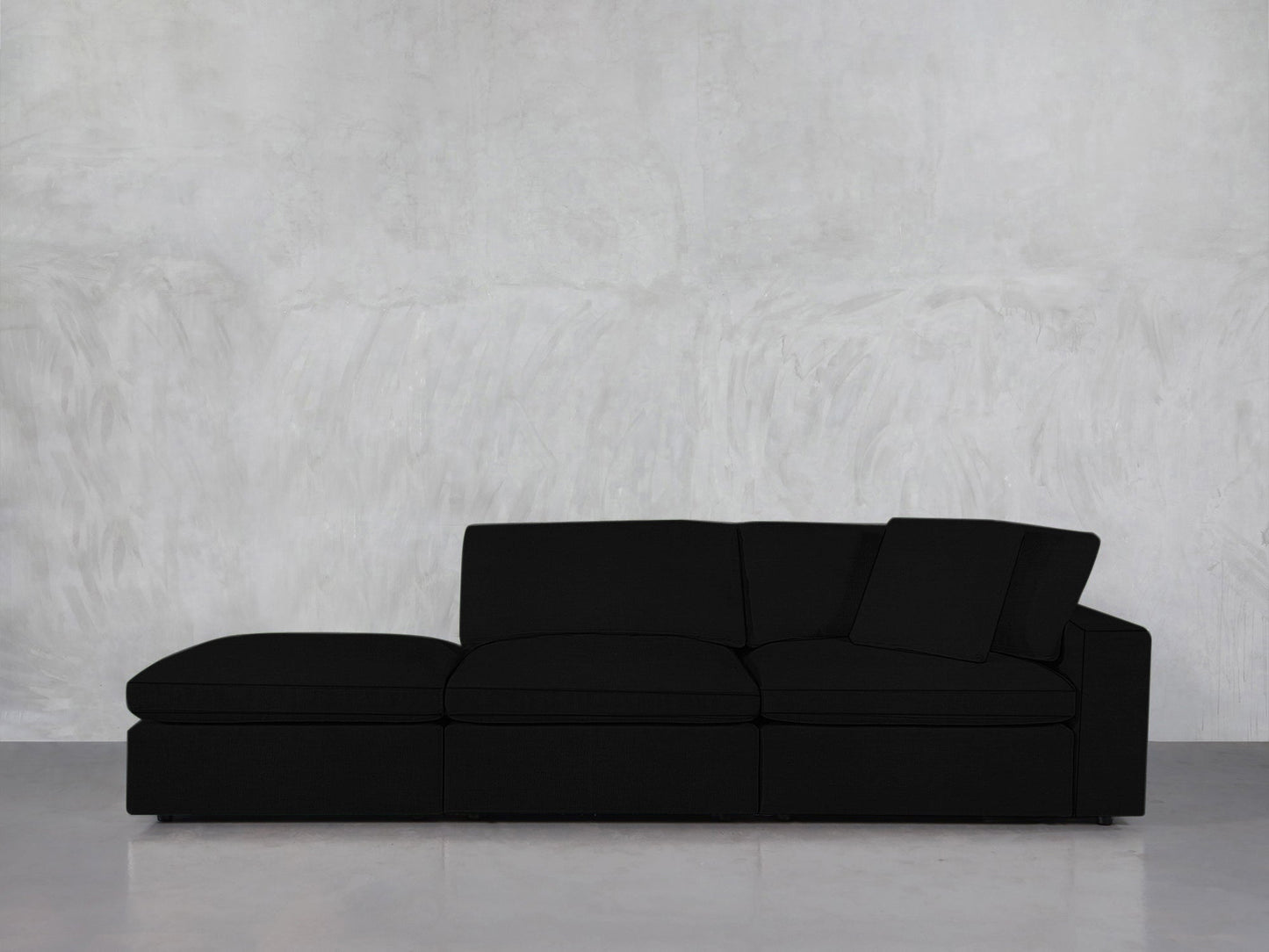 A 3-Seat Modular Lounger Sofa in modern black, paired with a matching cushion, is set against a light gray textured wall on a smooth, light-colored floor.