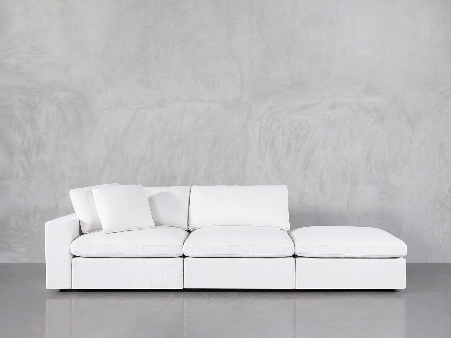 A 3-Seat Modular Lounger Sofa in white with three seat cushions and an ottoman is placed before a light gray textured wall and a reflective gray floor. One white pillow is positioned on the left side of the sofa.