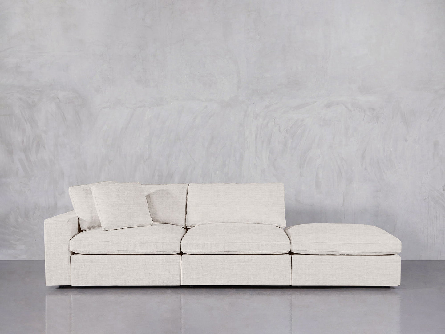 The 3-Seat Modular Lounger Sofa in light beige features three seats, a chaise lounge, and two matching cushions, displayed against a gray textured wall and reflective gray floor.