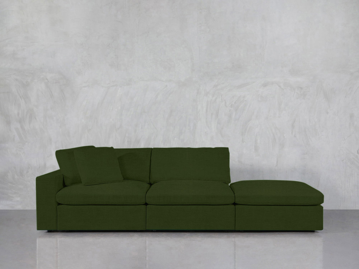 The 3-Seat Modular Lounger Sofa, featuring a modern green design with three seat cushions, one back cushion, and a chaise lounge, is positioned against a plain light gray textured wall on a smooth light-colored floor.