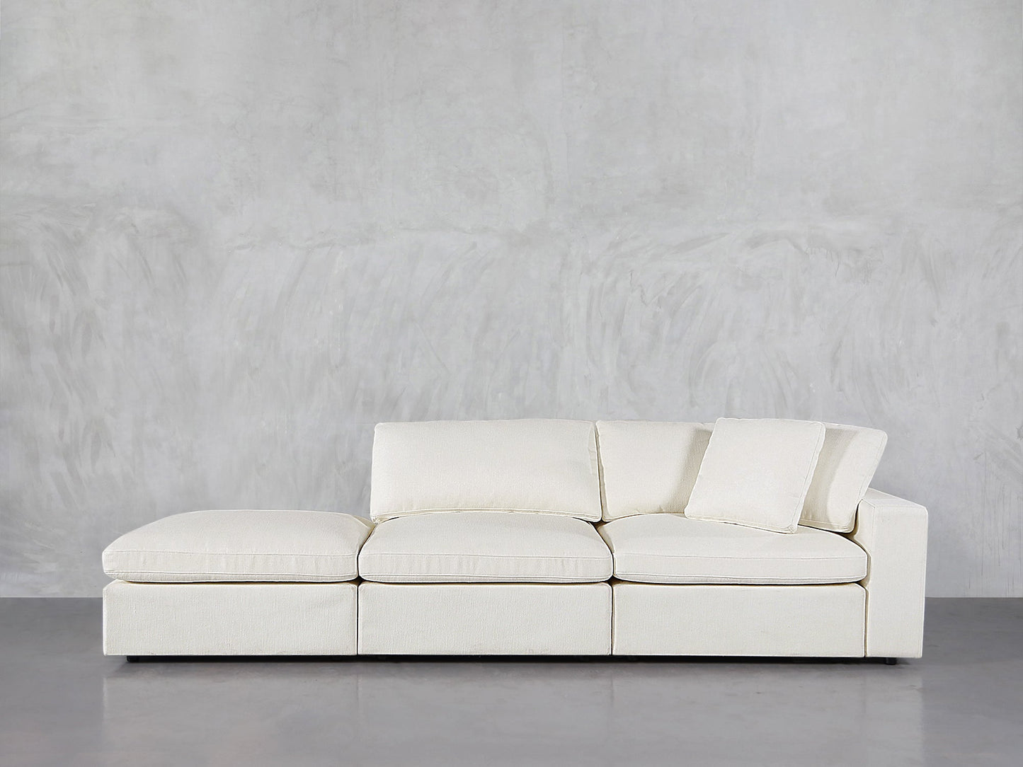 The 3-Seat Modular Lounger Sofa, in modern white with clean lines, sits against a light gray textured wall and polished concrete floor. One cushion is propped upright on the right side.