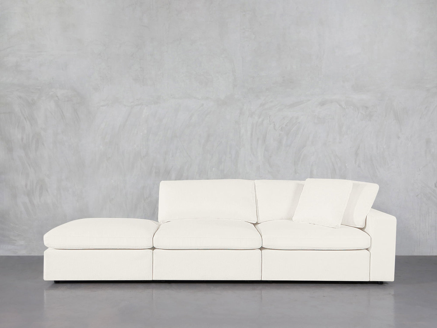 A 3-Seat Modular Lounger Sofa in modern white, accompanied by a matching pillow, is placed against a light gray textured wall on a smooth, reflective floor.