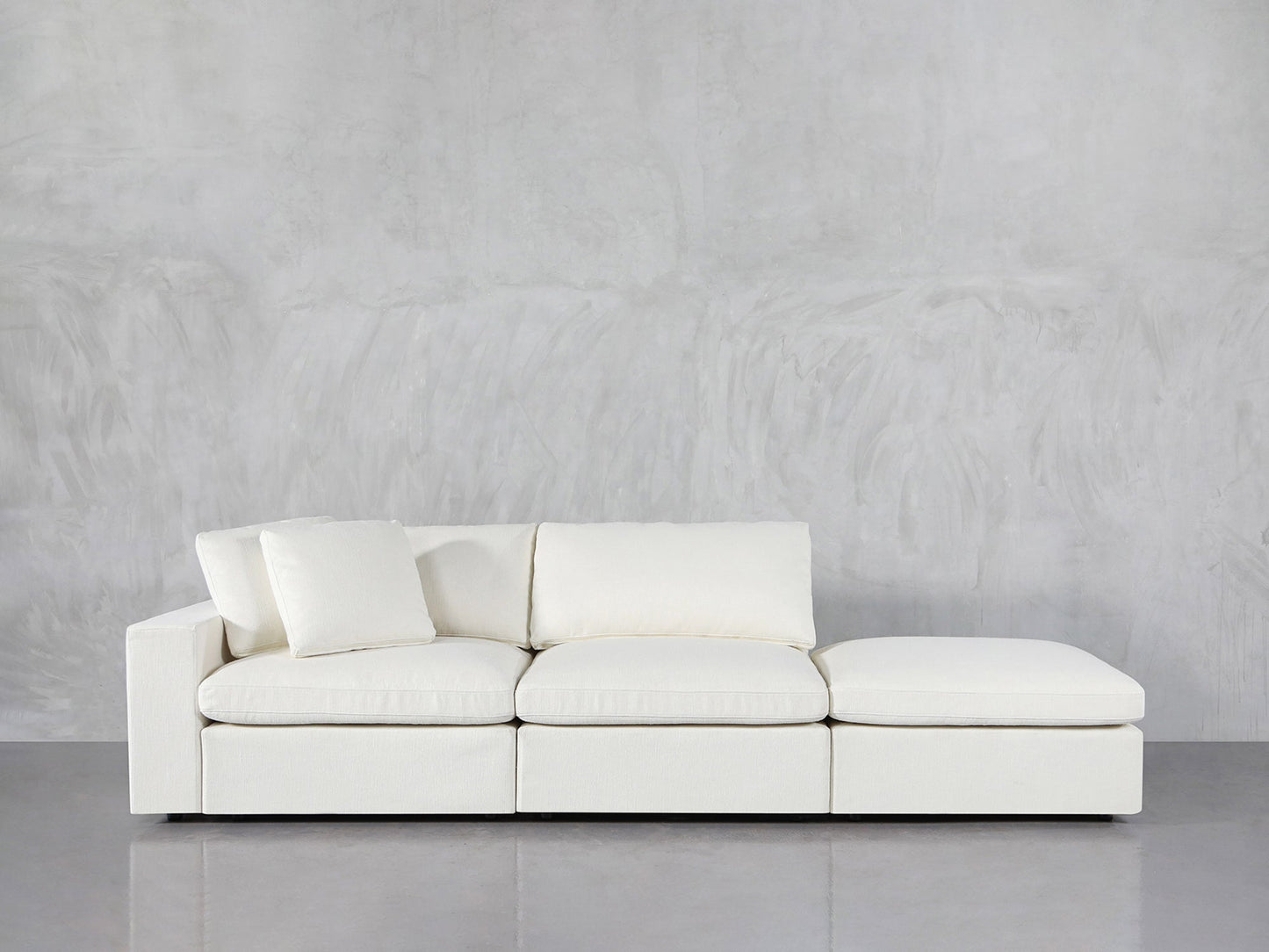 The 3-Seat Modular Lounger Sofa, featuring a modern white design with an attached chaise lounge, is placed against a light gray textured wall on a polished, reflective floor.