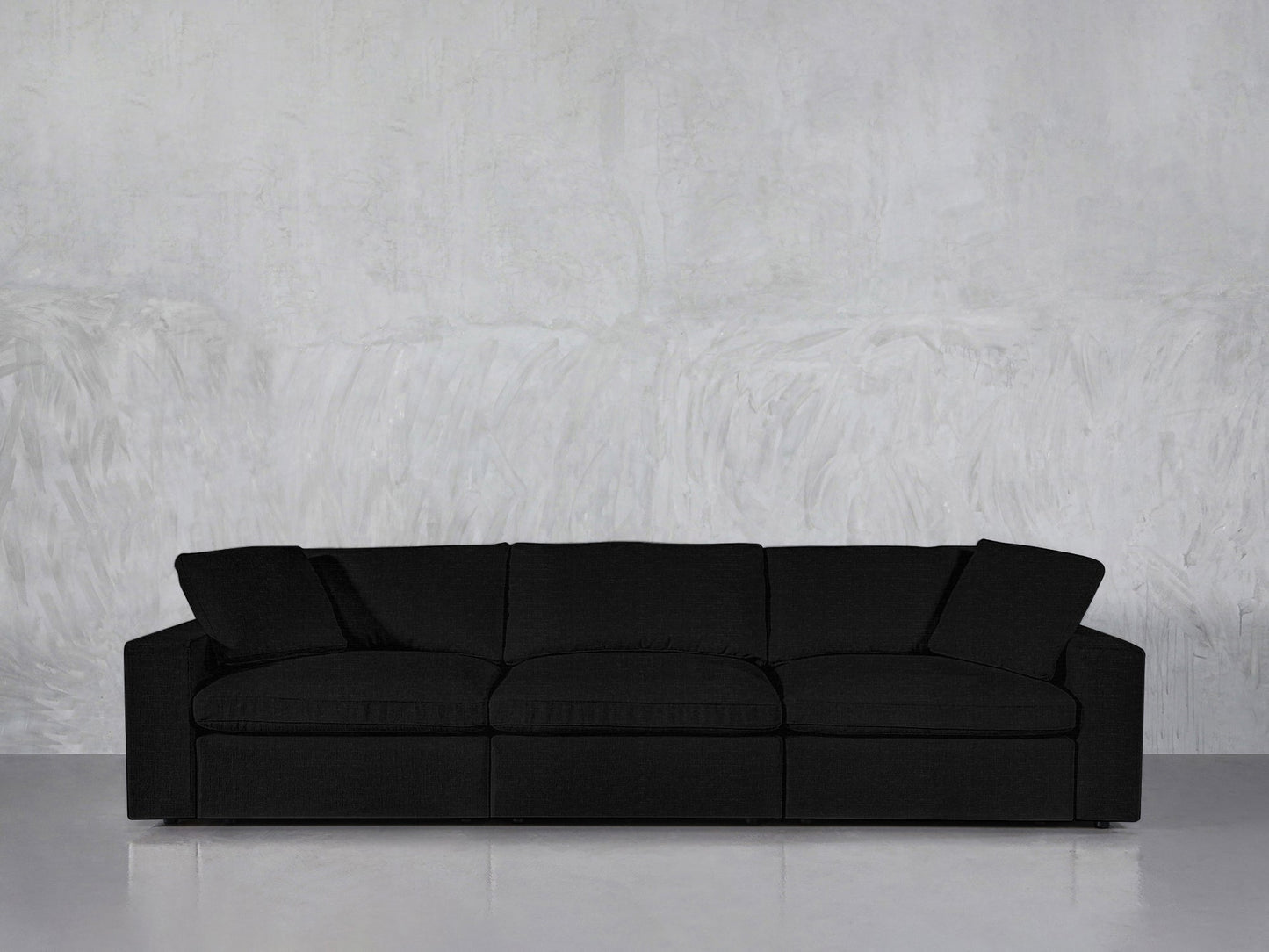 The 3-Seat Modular Sofa in modern black, featuring three seat cushions and two armrest pillows, is positioned against a light gray textured wall on a smooth, reflective floor.