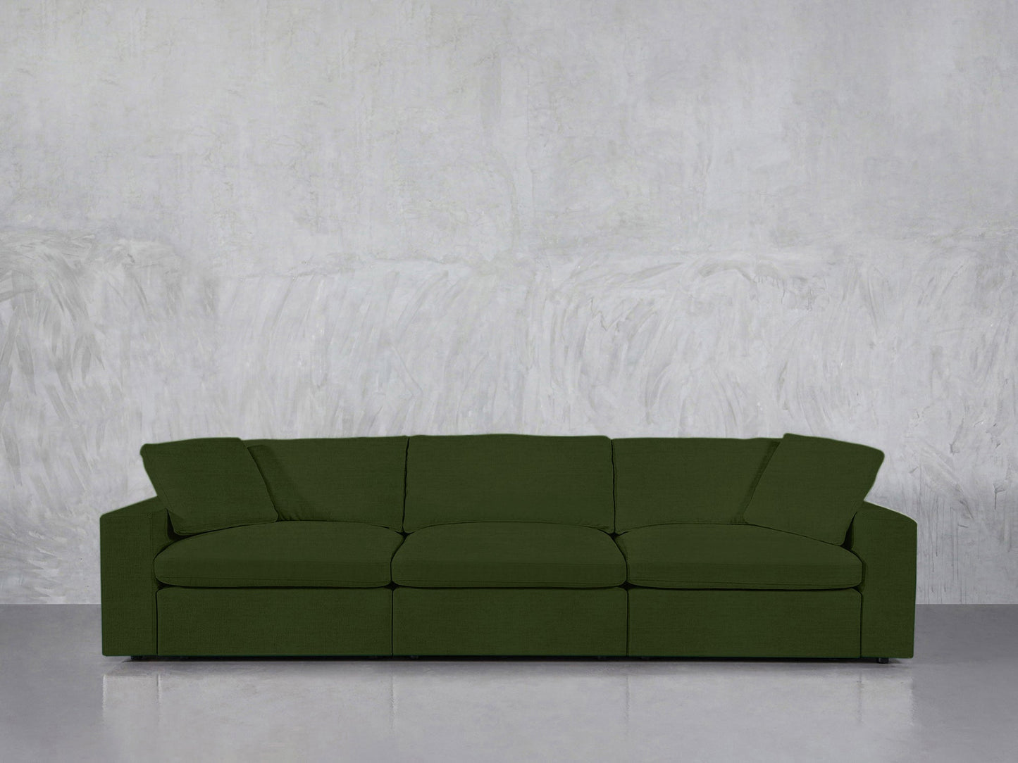 A 3-Seat Modular Sofa in dark green sits against a light gray textured wall on a shiny, light-colored floor. It features wide armrests and plush cushions.