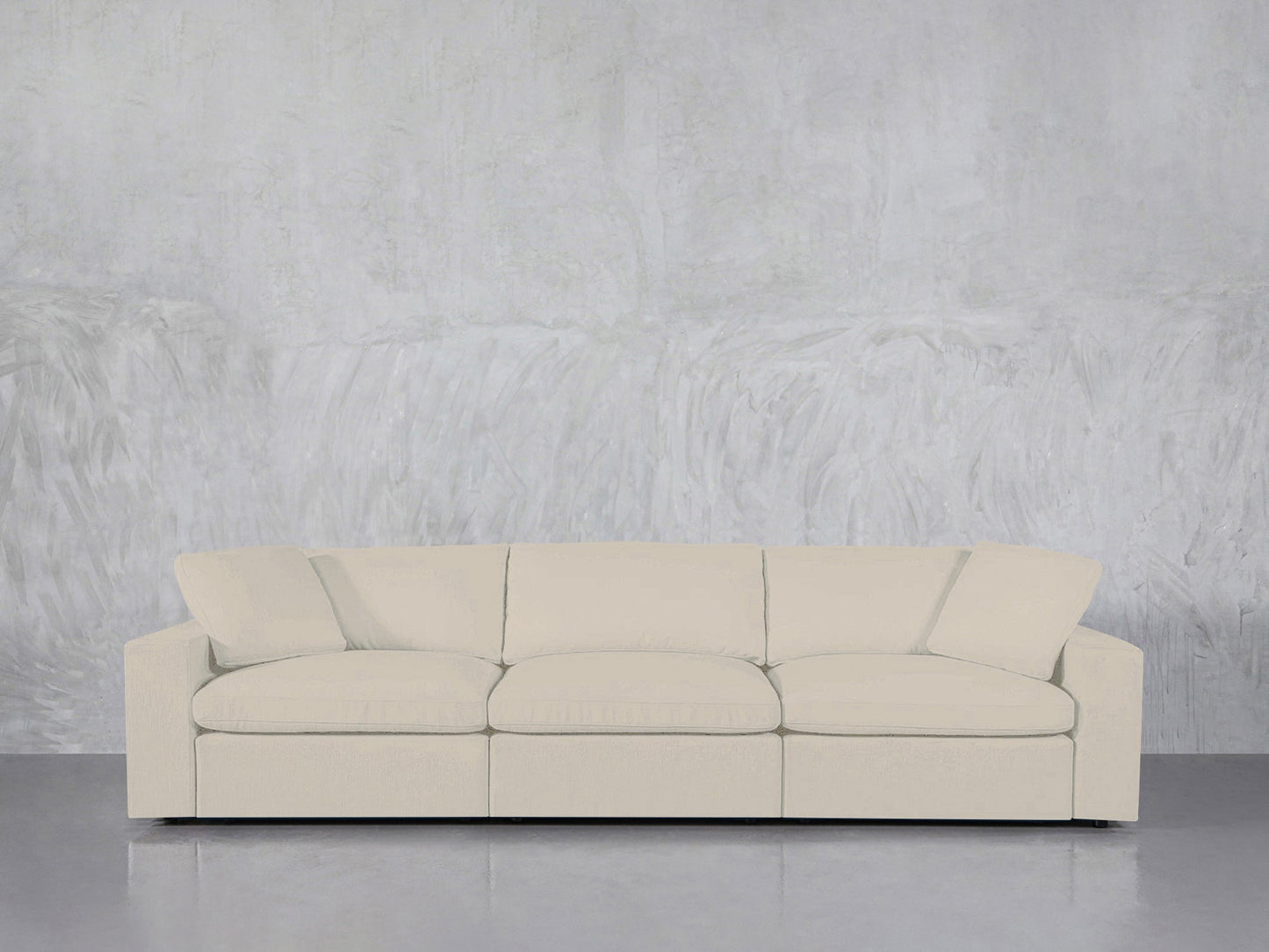 The 3-Seat Modular Sofa, in cream color with three seat cushions and an angled back cushion, is set against a textured light gray concrete wall and a smooth gray floor.