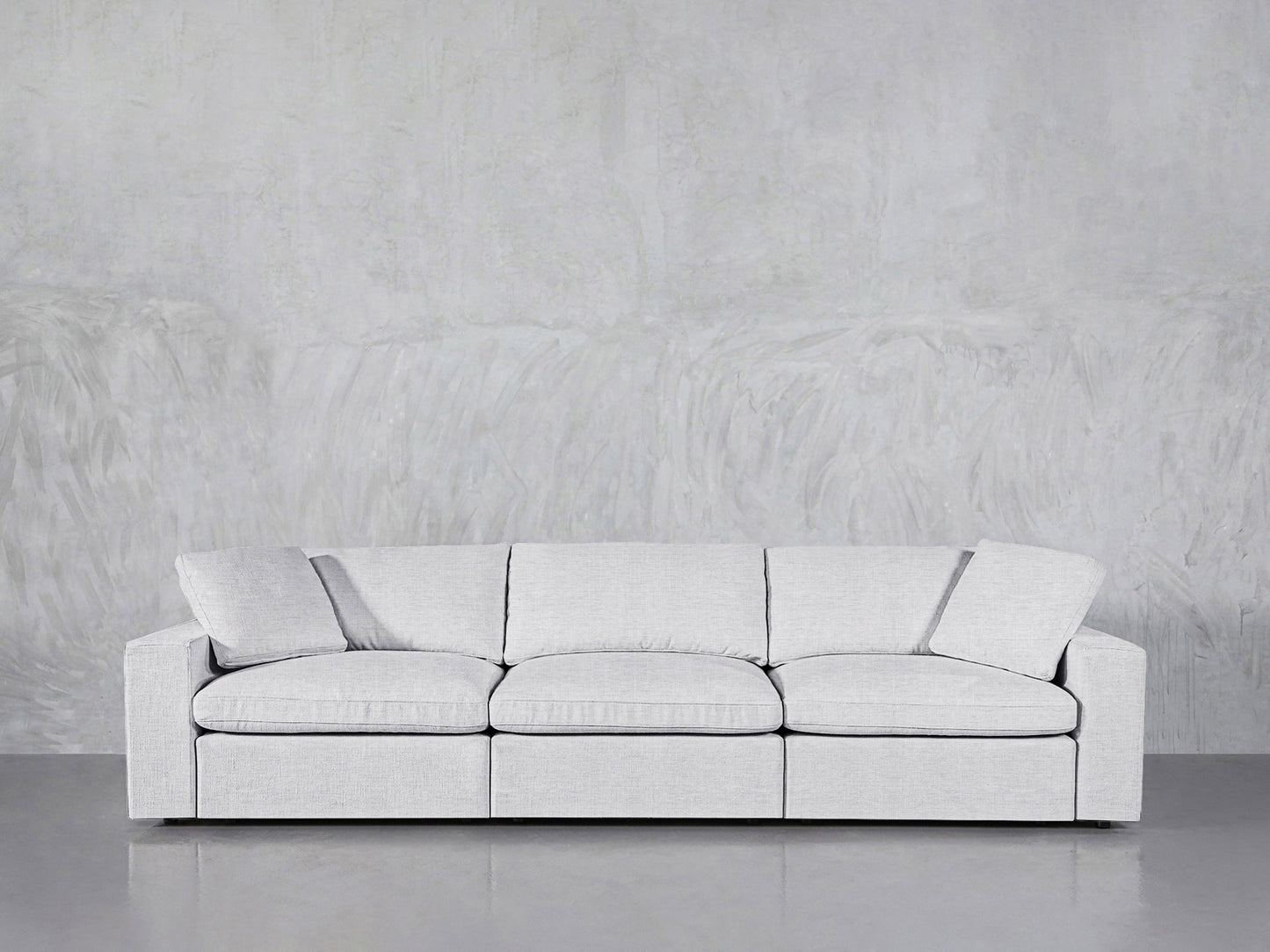 The 3-Seat Modular Sofa in light gray, featuring three seat cushions and a matching pillow, is placed against a textured gray wall and a smooth gray floor.