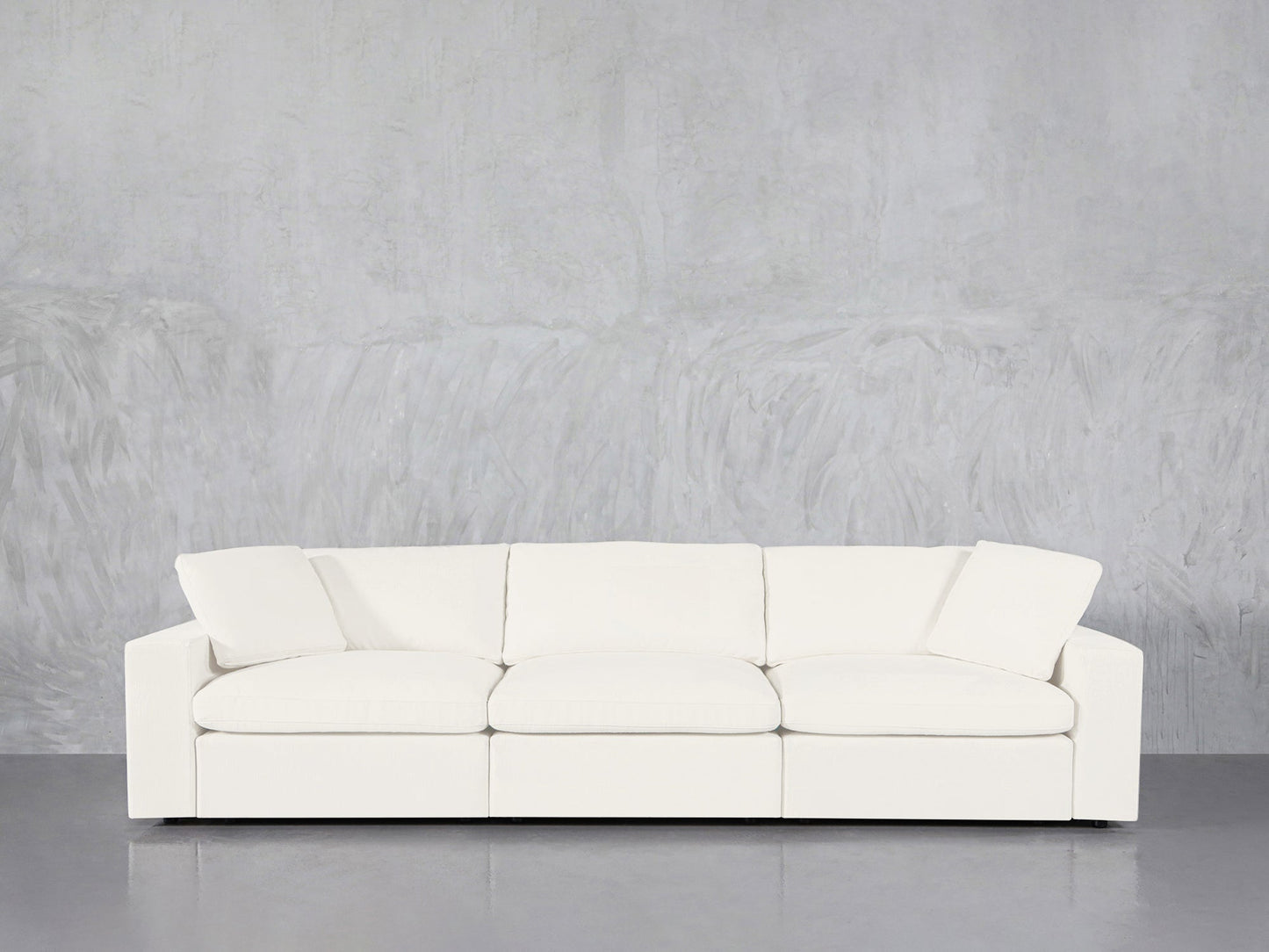 The 3-Seat Modular Sofa, featuring a modern white design with three seat cushions and two angled back pillows, is set against a light gray textured wall on a polished gray floor.