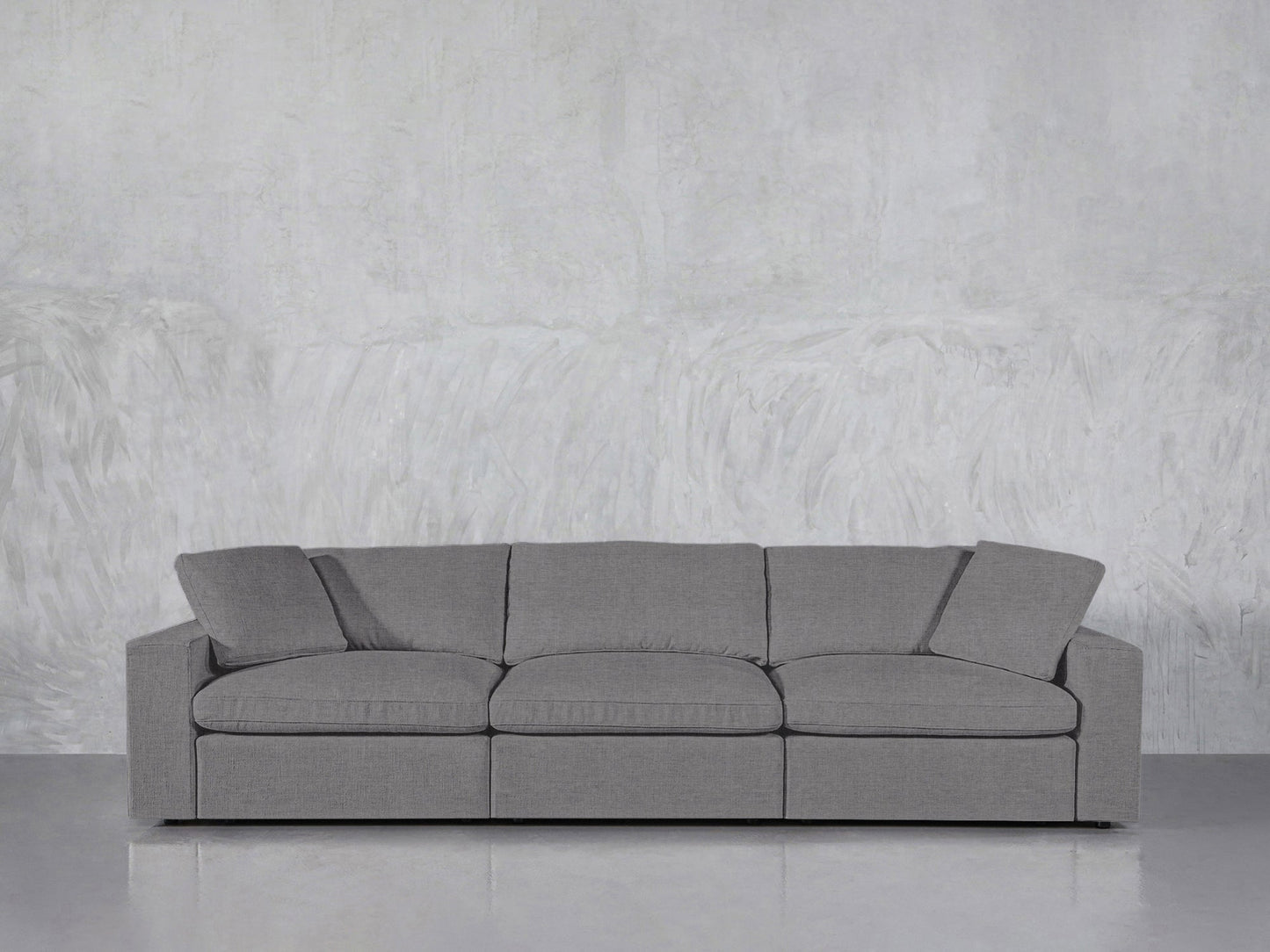 The 3-Seat Modular Sofa, upholstered in modern gray fabric with three seat and back cushions plus slightly tilted armrests, rests against a textured light gray wall on a smooth, light gray floor.