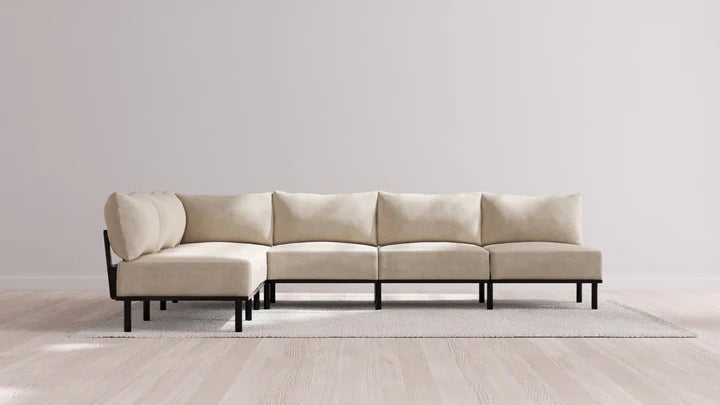 An Indoor Jack 6-Seater Open-Ends Corner Sectional in Balsa, featuring black legs, is elegantly positioned on a light gray rug against a plain white wall. The sectional is configured in an L-shape, enhancing the room's modern and minimalist appeal.