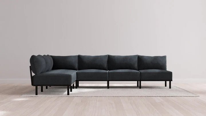 The Indoor Jack 6-Seater Open-Ends Corner Sectional in Balsa, with its sleek dark gray fabric, is styled in an L-shape on a light rug atop a wooden floor. The plain, light-colored wall behind complements the minimalist and contemporary aesthetic.