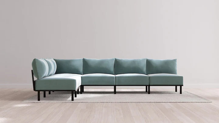 An Indoor Jack 6-Seater Open-Ends Corner Sectional in Balsa, featuring light blue cushions and a black metal frame, is arranged on a beige rug. The seating rests against a plain light gray wall on a light wood floor.