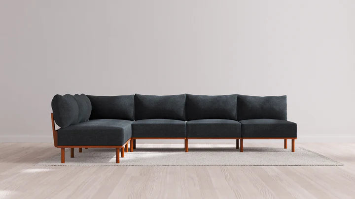 The Indoor Jack 6-Seater Open-Ends Corner Sectional in Balsa, with its modern design and dark gray upholstery, is elegantly positioned on a light-colored rug in a minimalist setting featuring light wood flooring and a simple, pale wall backdrop, complemented by its stylish wooden legs.