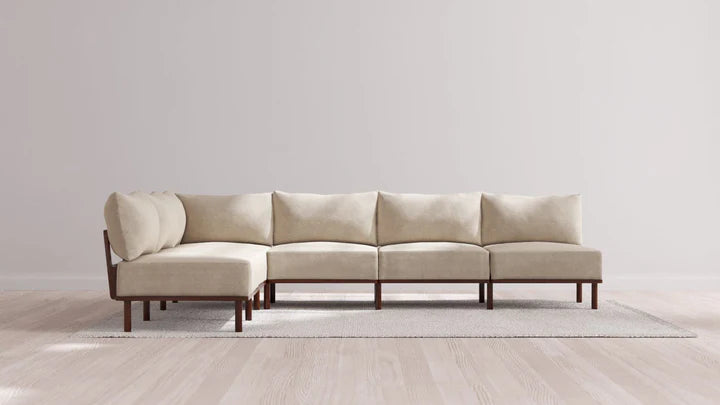 An Indoor Jack 6-Seater Open-Ends Corner Sectional in Balsa, featuring an L-shaped modular design and wooden legs, rests on a light gray rug within a minimalist room adorned with light wood flooring and a plain white wall.