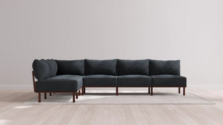A dark gray Indoor Jack 6-Seater Open-Ends Corner Sectional in Balsa with wooden legs sits on a light-colored area rug on a wooden floor. The background is a plain, light gray wall.