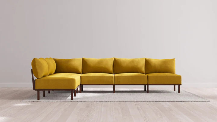 The Indoor Jack 6-Seater Open-Ends Corner Sectional in Balsa, featuring a vibrant yellow hue, sits on a light gray rug in a minimalist room. It boasts cushioned seats and backrests with wooden legs and is set against a plain white wall and a light wooden floor.