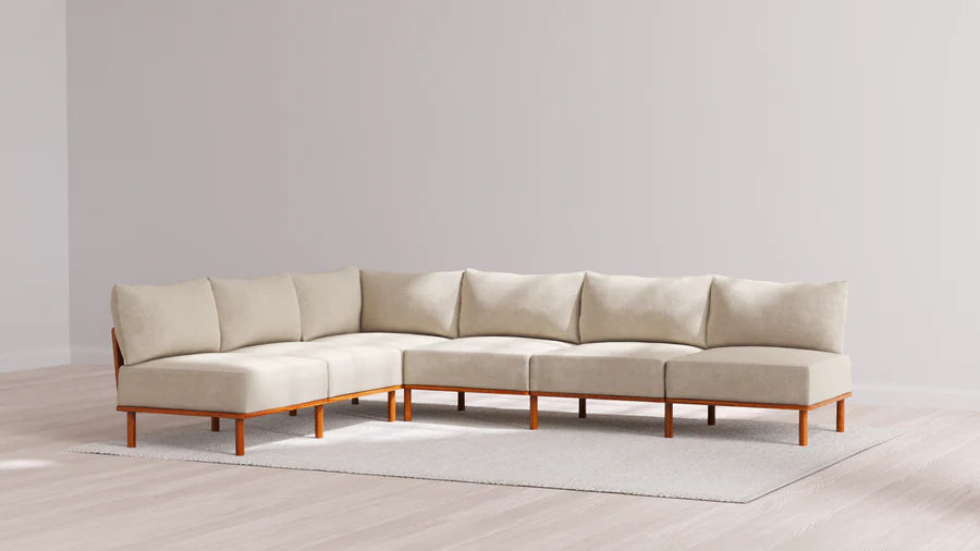 An Indoor Jack 6-Seater Open-Ends Corner Sectional in Balsa, featuring an L-shaped design with wooden legs, is set on a light gray rug in a minimalist room with wooden flooring and a plain light gray wall.