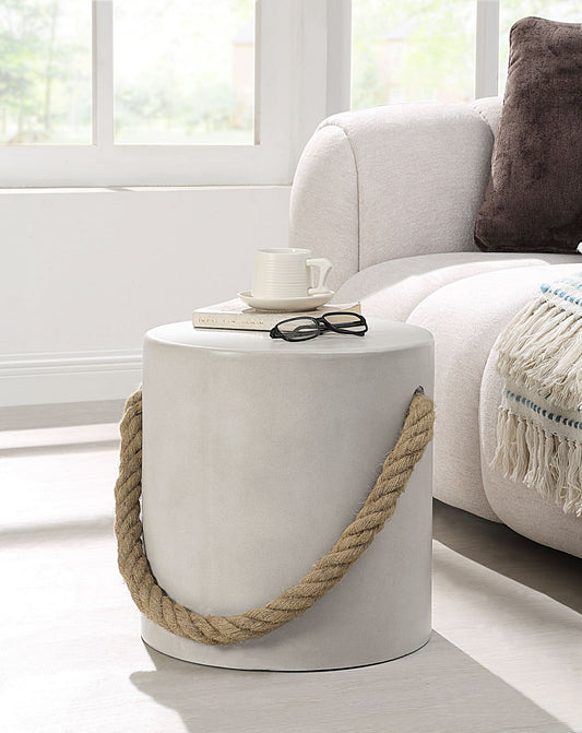 The Gene - Fiber Concrete Accent Table - Gray, featuring a thick rope handle, stands beside a beige sofa. On the table are a closed book, glasses, and a white cup and saucer. Soft natural light streams through large windows behind.