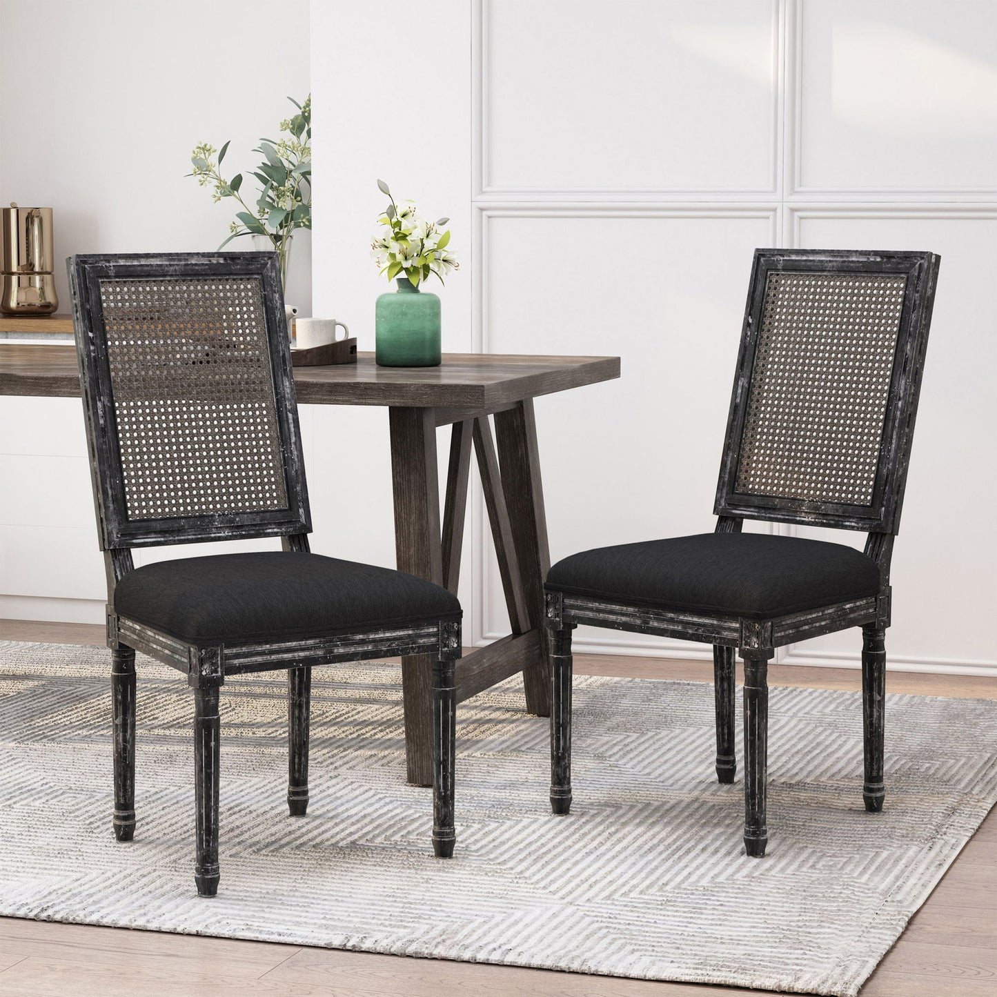 Two French Country Dining Chairs featuring rectangular cane backrests and smooth black upholstered seats are set on a light textured rug beside a dark wooden dining table, with decorative plants in the modern dining space.