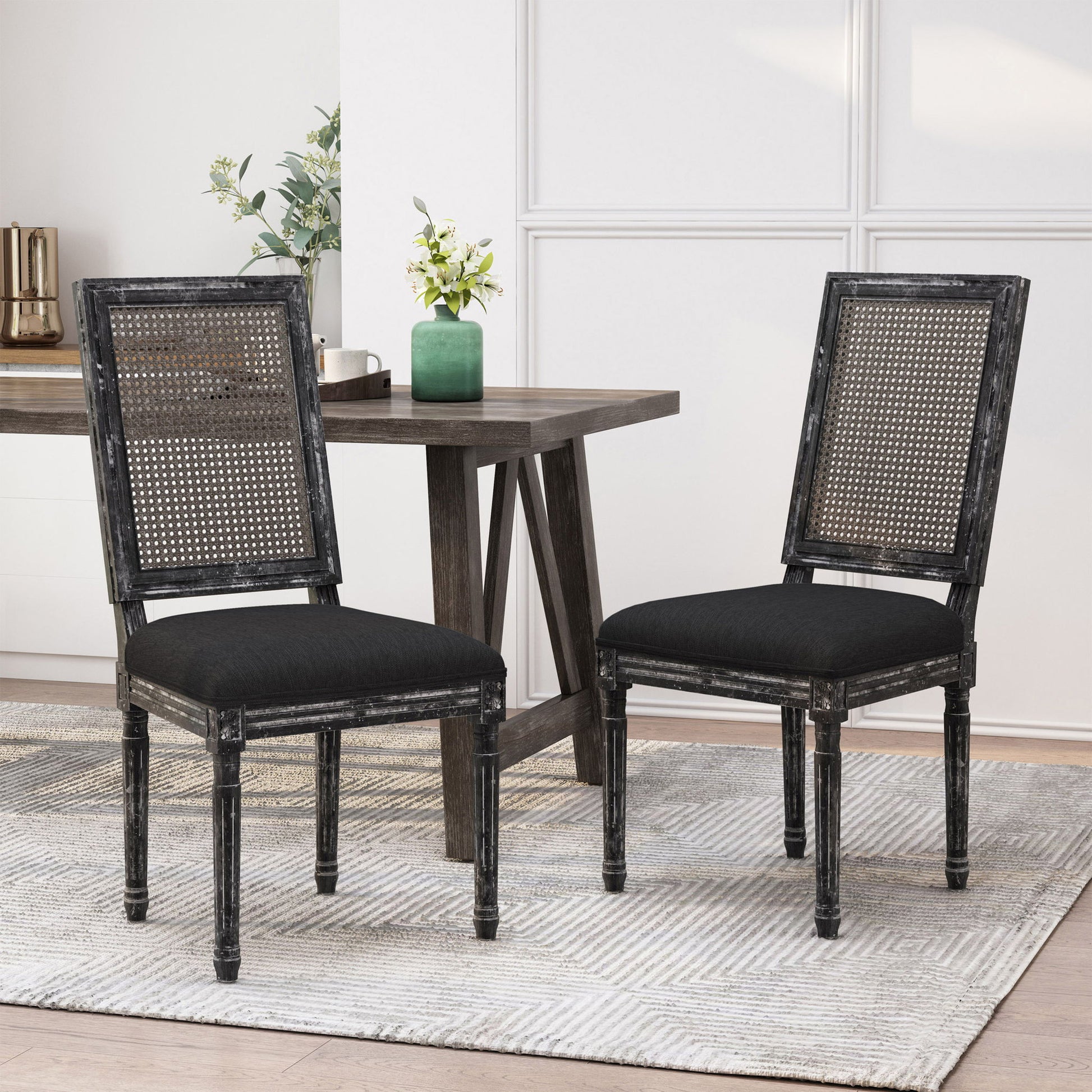 Two French Country Dining Chairs featuring rectangular cane backrests and smooth black upholstered seats are set on a light textured rug beside a dark wooden dining table, with decorative plants in the modern dining space.