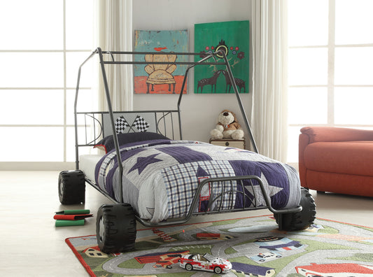 A children's bedroom with the Xander Twin Go Kart Bed in gunmetal, designed like an off-road car with large wheels and a metal frame. The bed is topped with a blue and white star quilt, surrounded by toys, books, and a teddy bear.