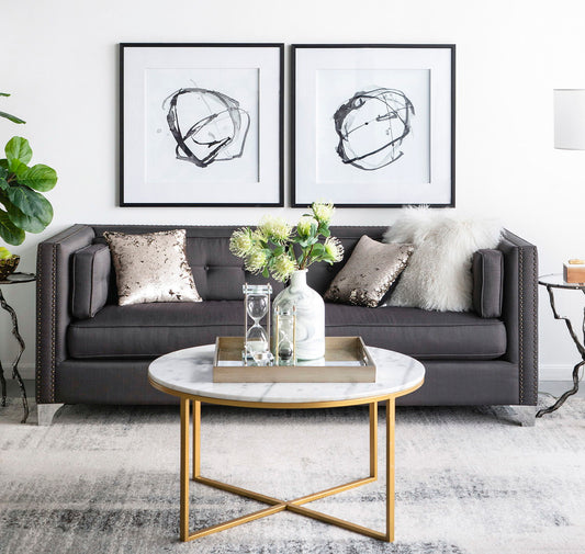 A modern living room with a dark gray sofa, metallic and fur pillows, a round marble coffee table with gold legs, Modern Abstract Wall Art (Set of 2) in black on the wall, plus decorative vases and plants.
