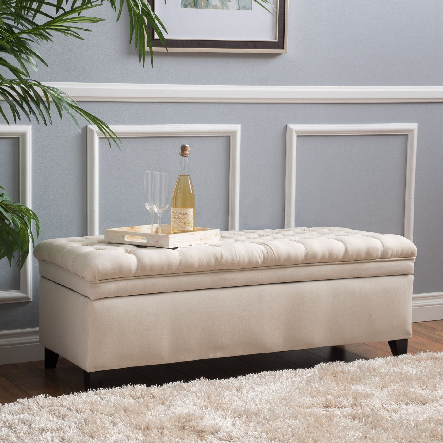 The Juliana Storage Ottoman with Birch Legs sits on a cream rug in the living room, topped with a wooden tray holding a bottle, two glasses, and a corkscrew. White paneled walls and a leafy plant complete the scene.
