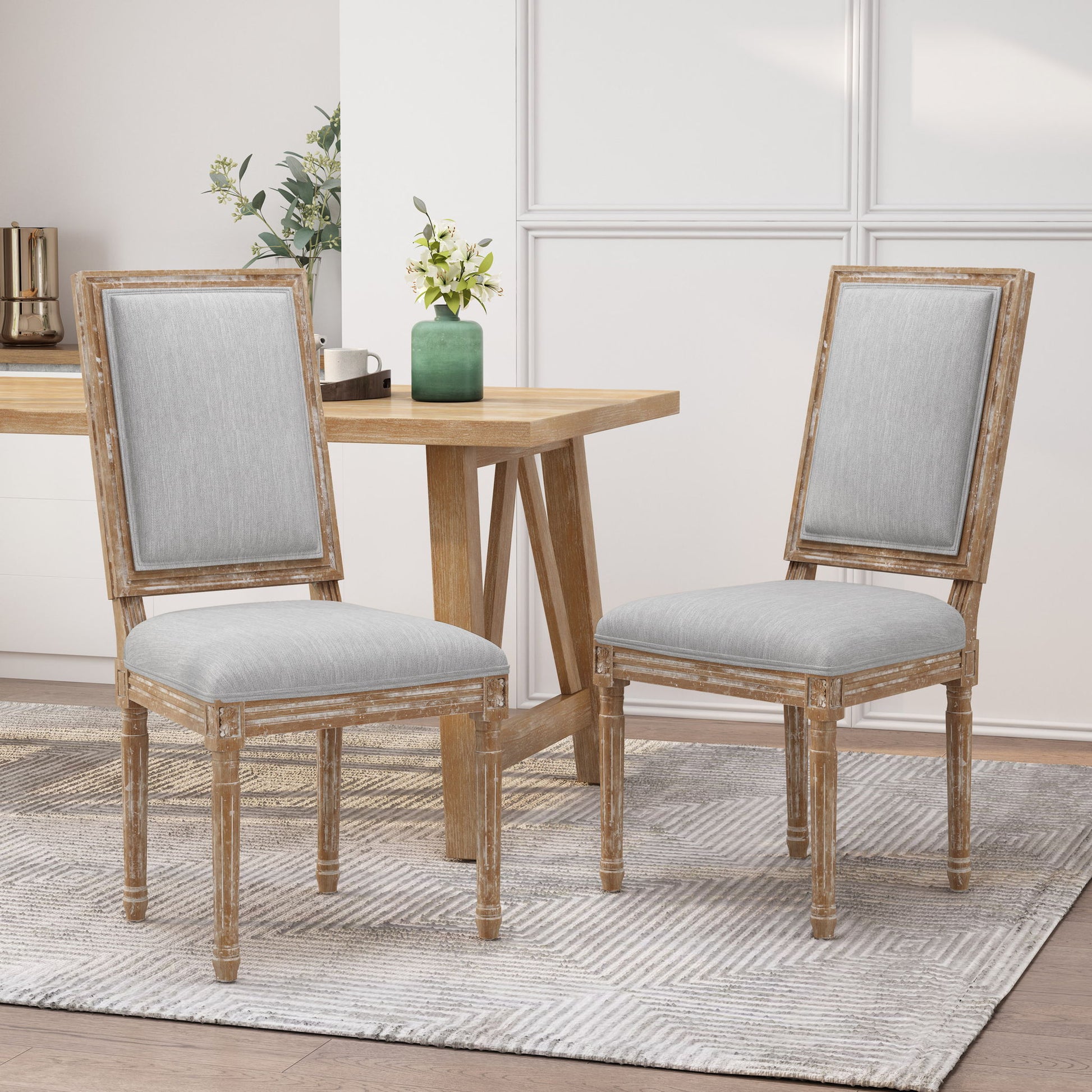 Two French Country Style Dining Chairs with upholstered seats and fluted legs sit on a patterned rug beside a light wood table in a bright, modern room with decorative plants and white paneled walls.