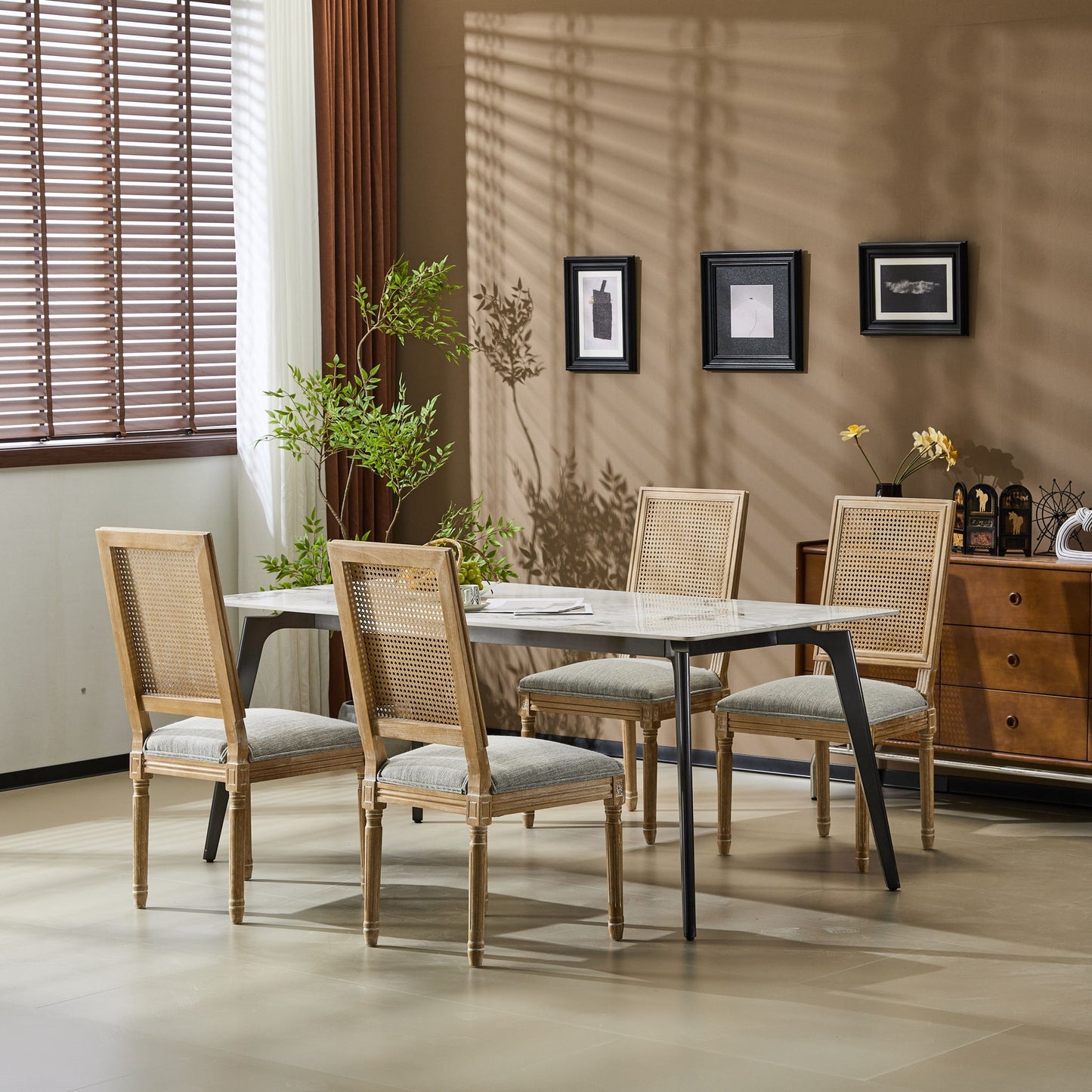 A dining room features a rectangular table, four French Country Dining Chairs with cane backrests and upholstered seats, a plant centerpiece, framed wall art, a decor-adorned sideboard, and sunlight streaming through window blinds.