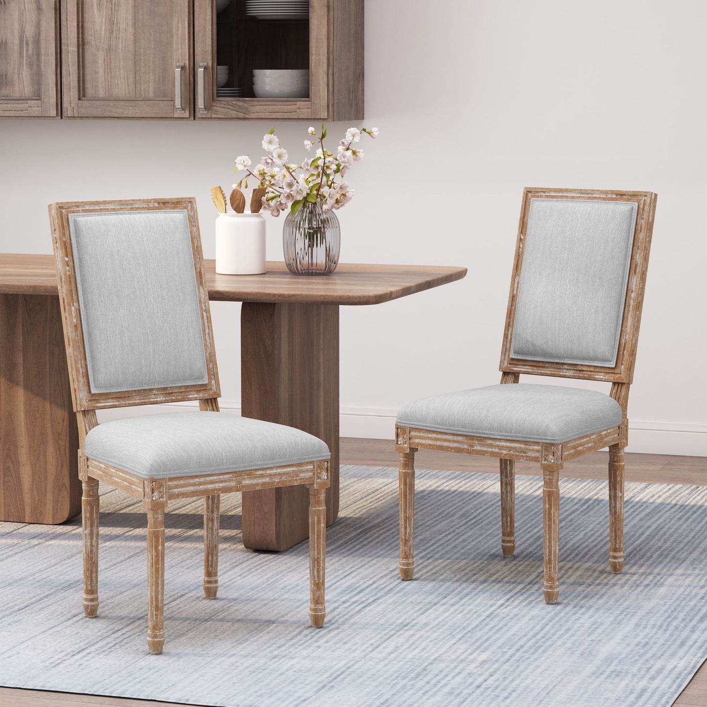 Two French Country Style Dining Chairs with upholstered seats and fluted legs sit on a blue-striped rug beside a wooden dining table topped with pink and white flowers in a modern dining space.