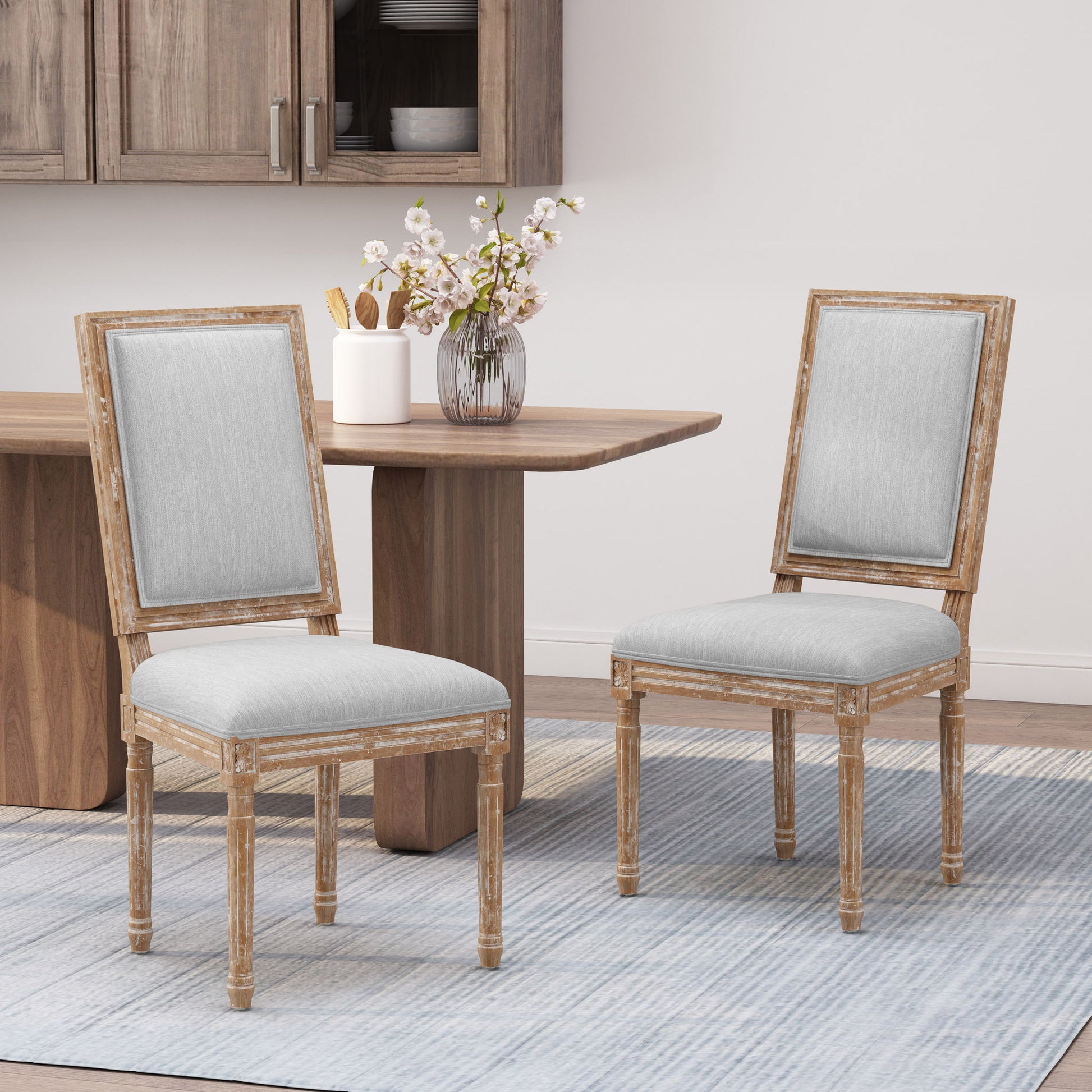 Two French Country Style Dining Chairs with upholstered seats and fluted legs sit on a blue-striped rug beside a wooden dining table topped with pink and white flowers in a modern dining space.