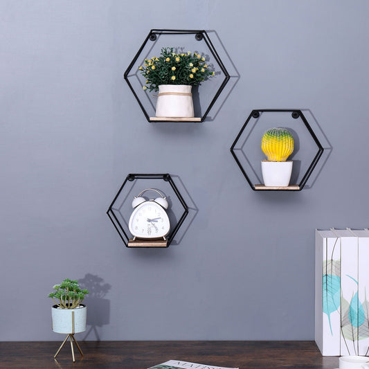 Three black Wall Mounted Hexagonal Floating Shelves (set of 3, natural wood) display two potted plants and a white alarm clock on a gray wall. Below, a small plant and books rest on wood. The look is minimalistic and modern.