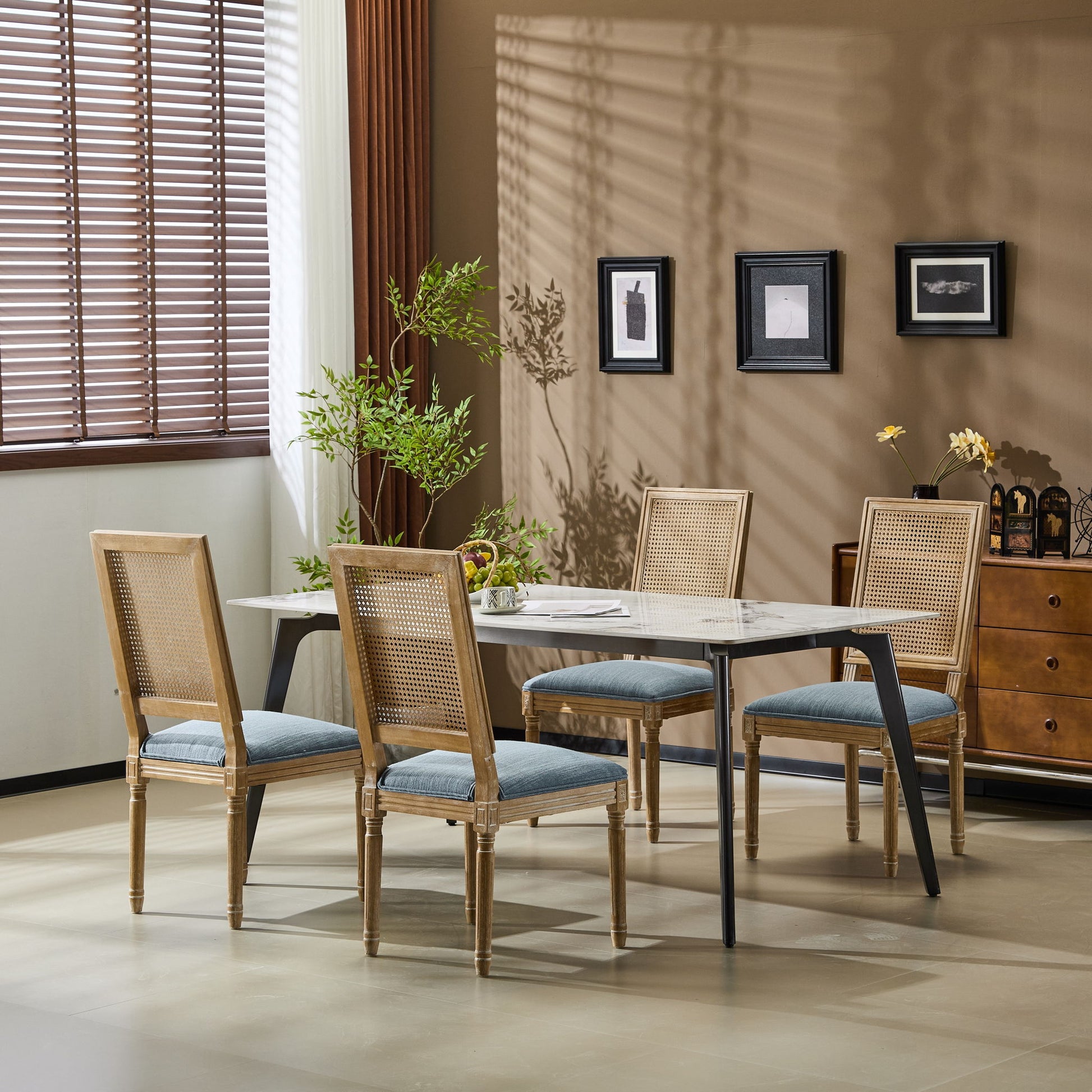 A dining room with a rectangular table, four French Country Dining Chairs featuring cane backrests and smooth upholstered seating, a potted plant, brown side cabinet, framed wall art, and sunlight streaming through window blinds.