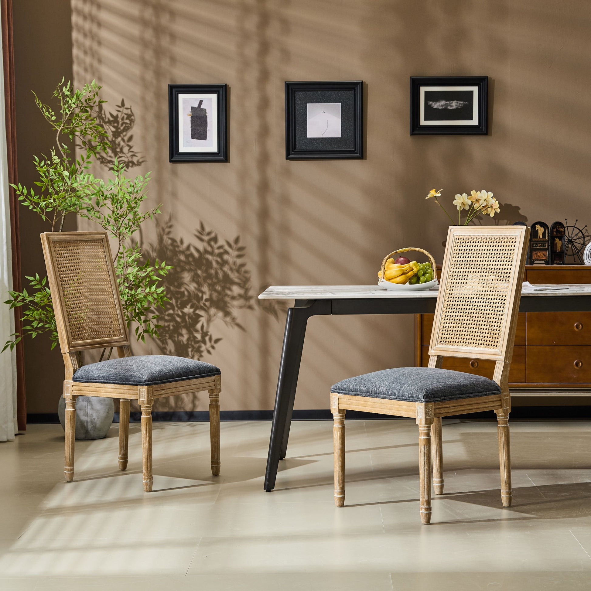 A modern dining room featuring two French Country Dining Chairs with rectangular cane backrests and smooth upholstered seating, a rectangular table with a fruit bowl, wall art, sideboard, potted plants, and sunlight casting shadows on beige walls and floor.