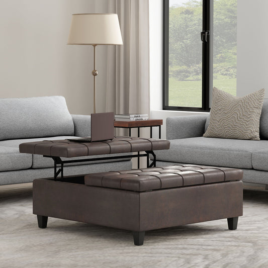 The Harrison Large Coffee Table Storage Ottoman, in brown and square-shaped with a lift-top tray, is placed between two gray sofas in the living room. A lamp and book are on a side table, and a large window fills the space with natural light.