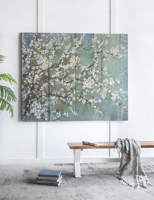 A set of four Saison Cherry Blossom Canvas Prints featuring white blossoms and teal backgrounds decorates a white wall above a wooden bench with a gray throw and books; a green plant is partially visible on the left.