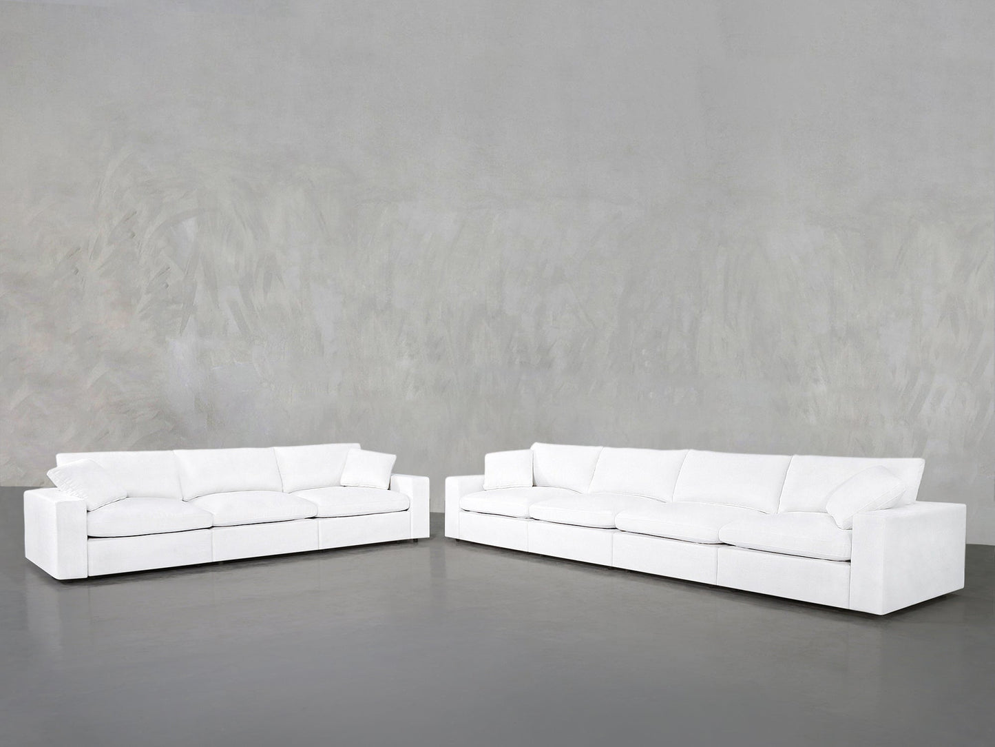 The 4-3 Sofa Set, featuring two modern white sofas with plush cushions, is arranged in an L-shape against a light gray wall and dark gray floor for a minimalistic, spacious look.