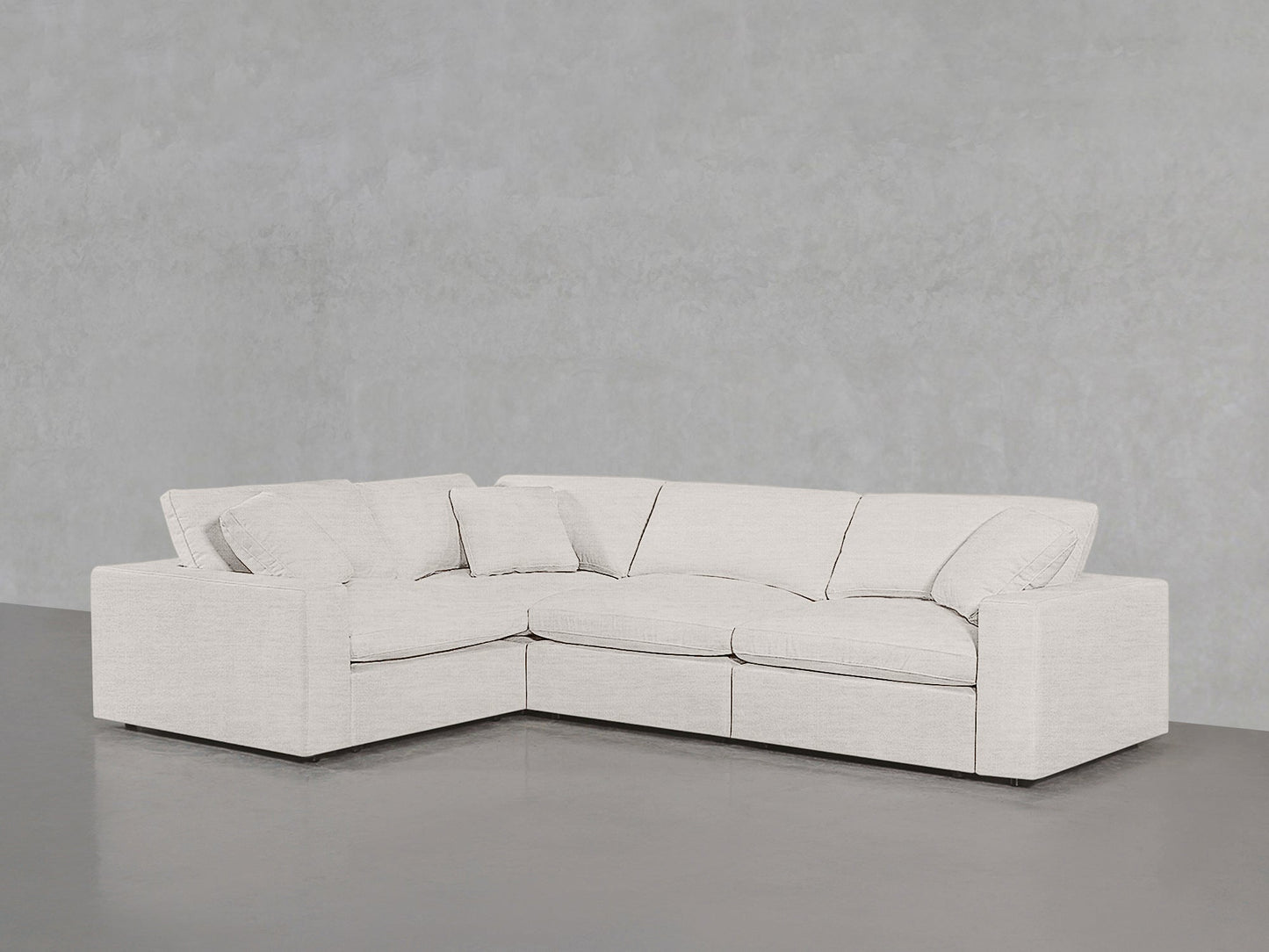 A modern 4-Seat Modular Corner Sectional in light gray with matching pillows sits against a plain light gray wall on a smooth gray floor.
